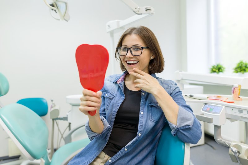 a middle-aged woman wearing glasses looking at her new smile in the mirror at the dentist’s office thanks to dental implants