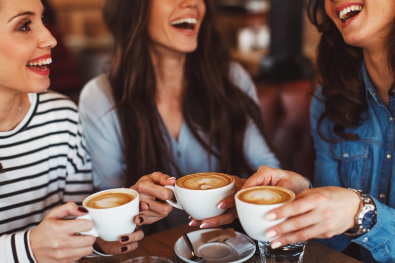 Three women at a small wooden table laughing over hot lattes in white mugs