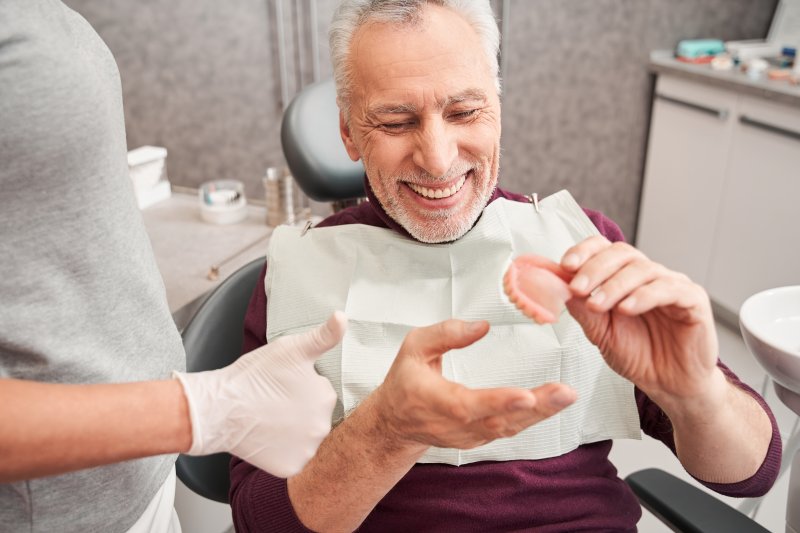 Elderly man smiling in dental chair holding dentures as dentist stands to the side with a thumbs up