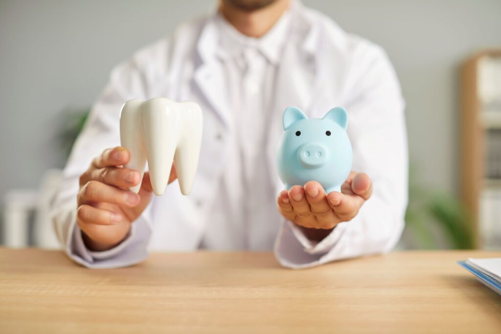 Neck-to-chest view of dentist in white coat holding model tooth and piggy bank